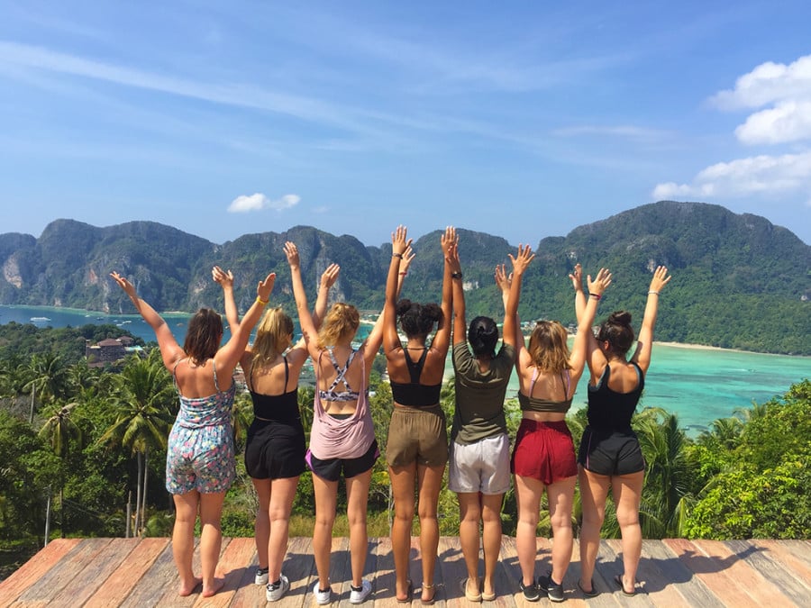 Travellers posing with their hands in the air in front of a blue sea and lush greenery