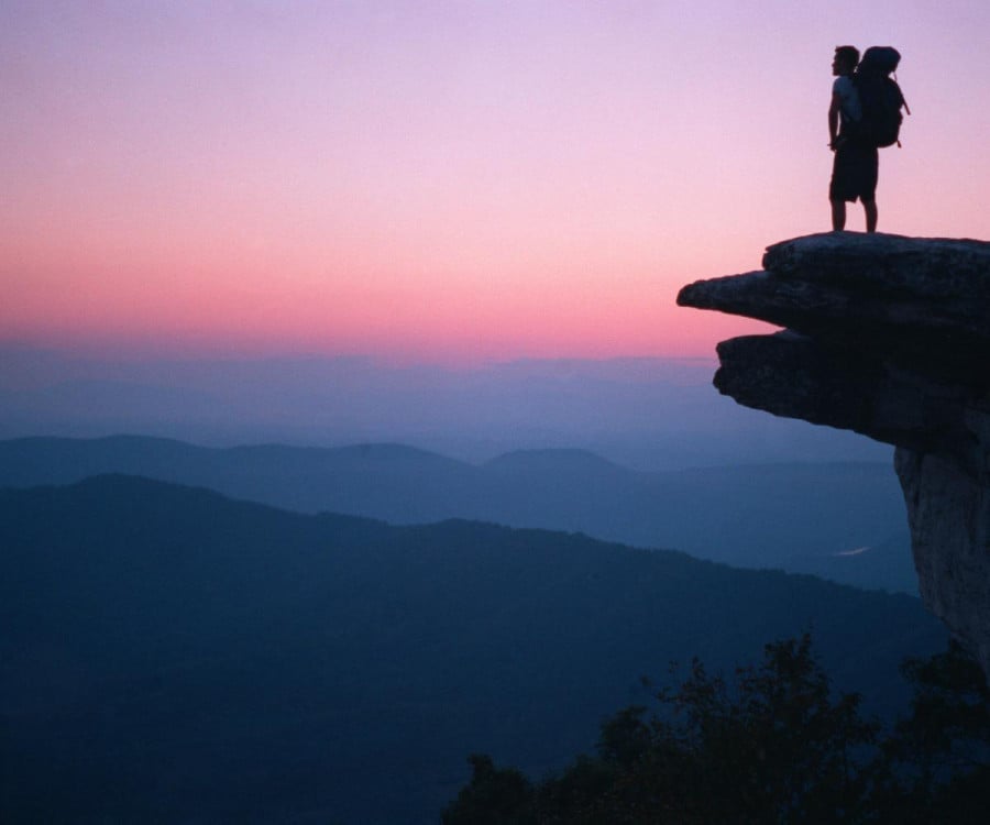 A traveller with a backpack standing on top of a peak overlooking mountains at sunset
