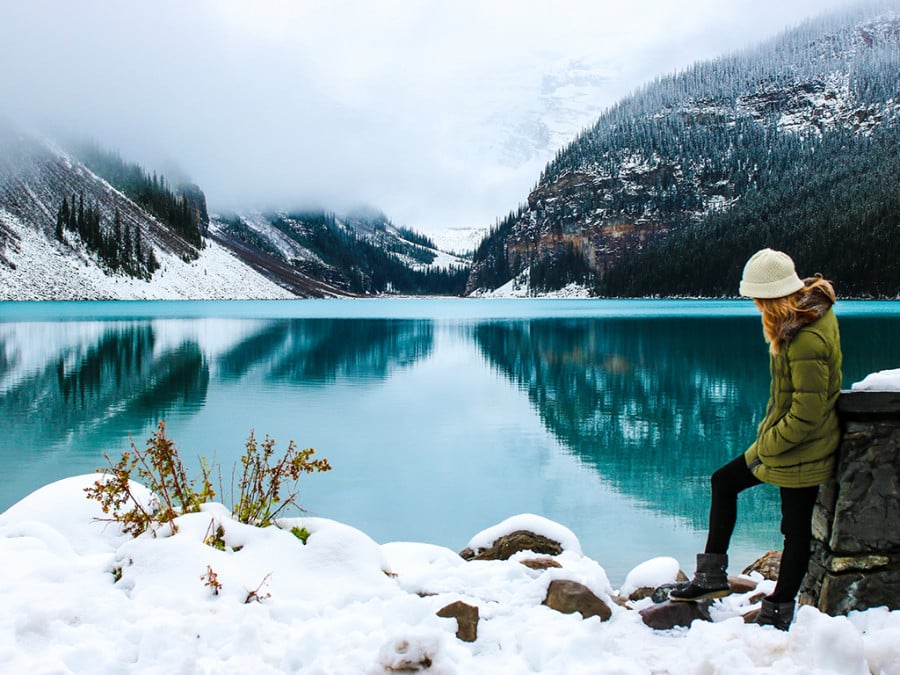 A person standing on snow in front of a bright blue lake and pine forested mountains