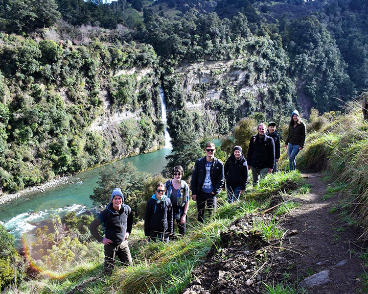 A group of people standing on a path along River Valley, New Zealand with a waterfall and river