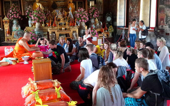 A group of travellers sit in a temple with one receiveing a blessing from a Buddhist monk