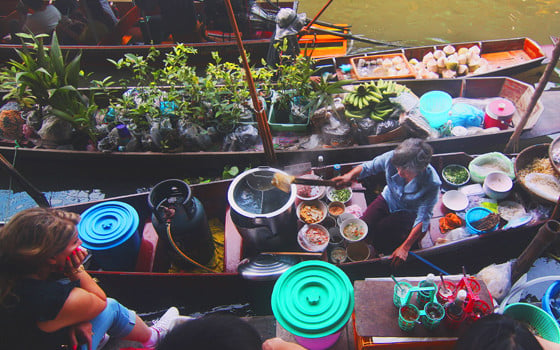 A traveller looking at a person cooking food in a boat at a floating market